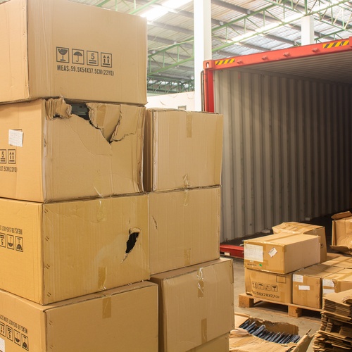 Multiple pallets of damaged cardboard shipping boxes sit next to an unloaded freight truck near a loading dock.