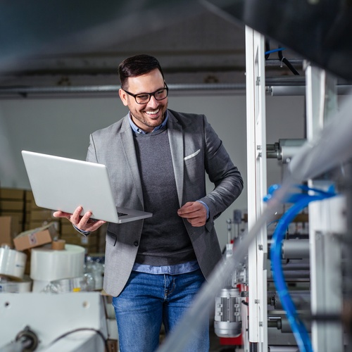 A man wearing a blazer holds a laptop in one hand and smiles while he walks through a room full of business equipment.