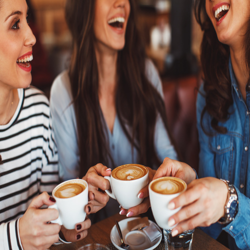 Three women enjoy coffee at a coffee shop together. They're all smiling at each other.