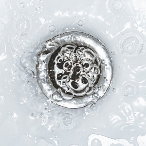 A puddle of water in a pristine, white sink slowly draining down a silver sink drain, creating bubbles.