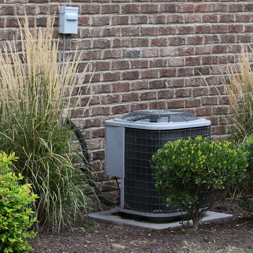 The outdoor half of a central AC unit sits on a concrete slab beside a few shrubs, ornamental grass, and a brick wall.