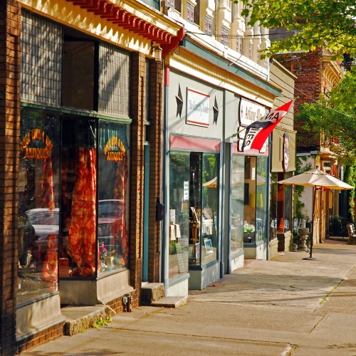 Small businesses line a sunny town street with a shared walkway in front. A flag hangs from one of the storefronts.