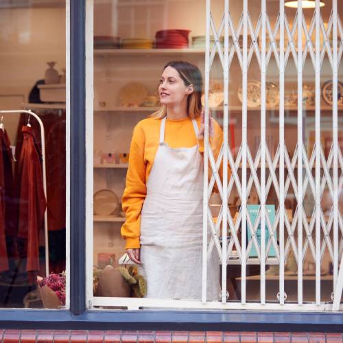 A female small business owner smiles as she slides open the security gate on the window of her clothing store.