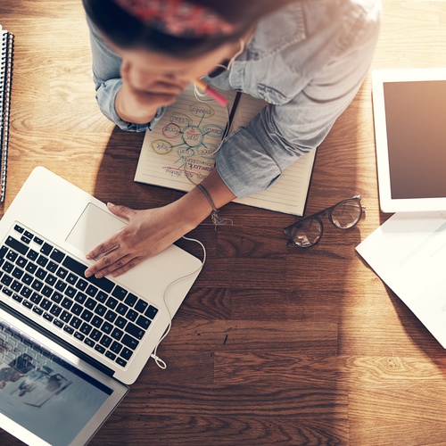 A top-down view of a woman working at a desk with a laptop, tablet, notebooks, and papers scattered about.