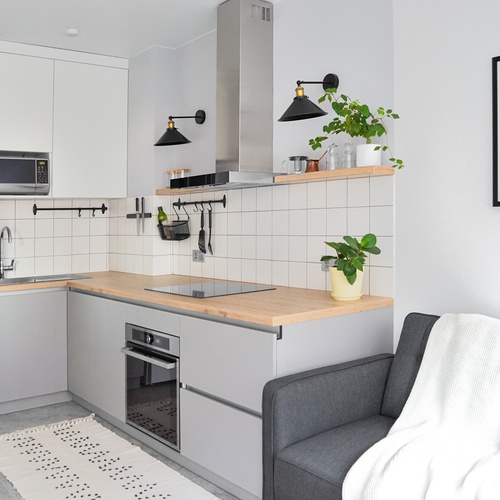 A small, grey kitchen with light wooden countertops, with hooks on the tile backsplash. In the corner is the grey living room couch.