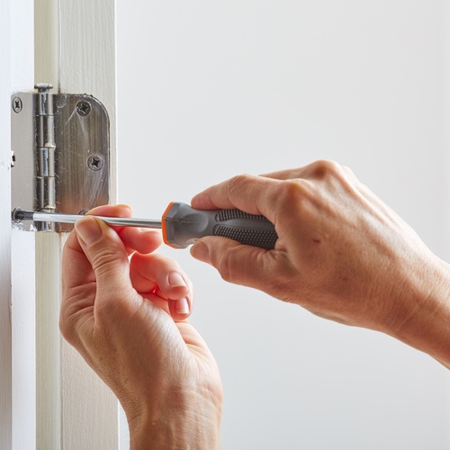 A pair of hands uses a gray-and-orange screwdriver to secure a silver door hinge on the doorframe.