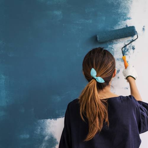 A woman with a blue bow in her ponytail uses a paint roller to paint a white wall a dark greenish blue color.