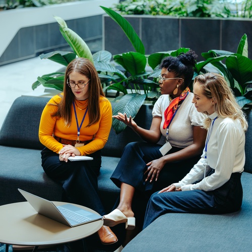Three women are sitting on a long, curved couch in a building with greenery behind them. They are looking at a laptop.