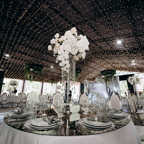 An elegant, sophisticated wedding reception with reflective table tops, white flowers, and string lights over the ceiling.