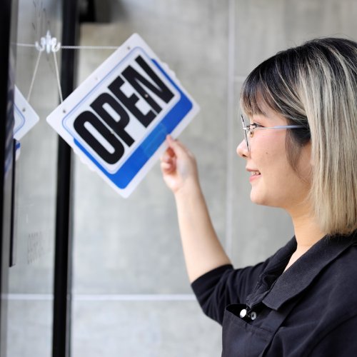 A woman with a short black and blonde bob haircut and a black apron flips the sign on a door to open with a smile.