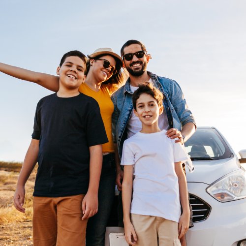 A family of four standing together in front of a car and smiling. Behind them is a road and a desert environment.