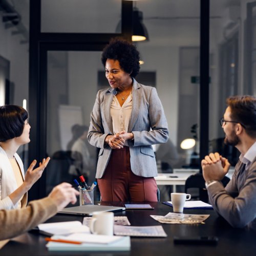 A woman wearing a gray suit smiles as she listens to another woman while conducting a group office meeting.