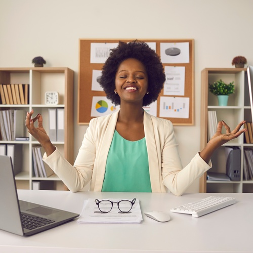 A Black woman sits at an office desk with her hands in a meditative position. She is leaning back in her chair and smiling.
