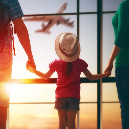 A family of three holds hands while looking out a large airport window at an airplane, with sunlight streaming through.