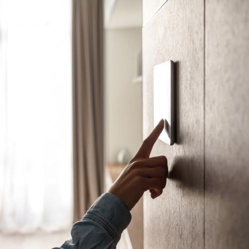 A woman smiling as she reaches out to press a button on her smart thermostat. She has one finger on the screen.