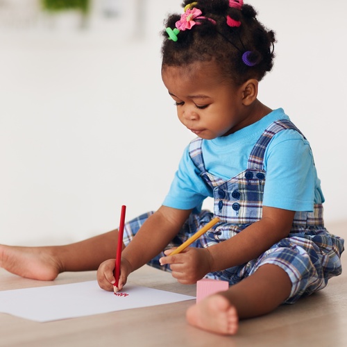 A toddler baby girl with colorful bows in her hair is concentrating and drawing with pencils using both hands.