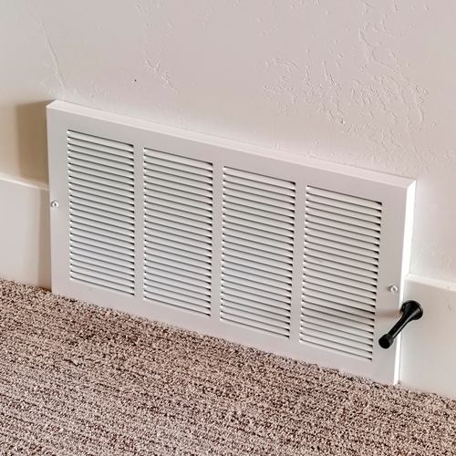 A close-up of a vent installed at the base of a wall that meets a brown carpet. The vent has a white plastic grille.