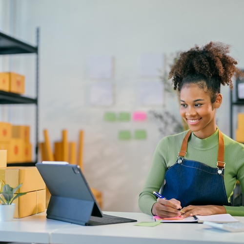 A young business owner writes on a notepad while looking at her tablet device. She smiles and sits at a desk.