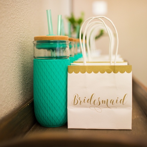 The top of a dresser with a set of matching teal straw cups and white bags labeled "bridesmaid" in gold text.