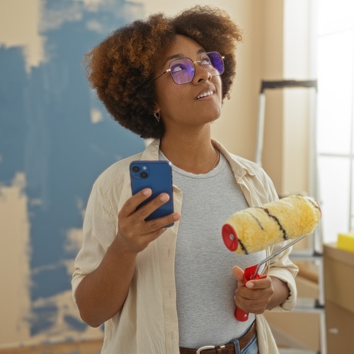 A young Black woman holds a phone and paint roller while looking up in a room with a ladder and partially painted walls.