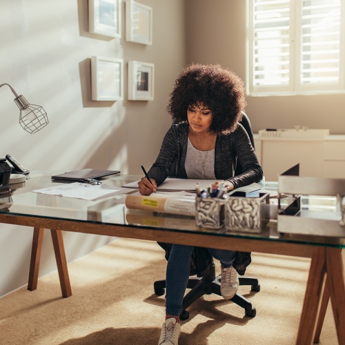 A woman sits at a wooden desk writing on a piece of paper. The desk also has a printer, a lamp, and a pen organizer.