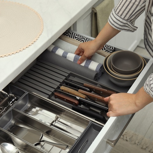 A drawer contains organized utensils, knives, stacked bowls, and kitchen towels. A person adjusts a folded towel.