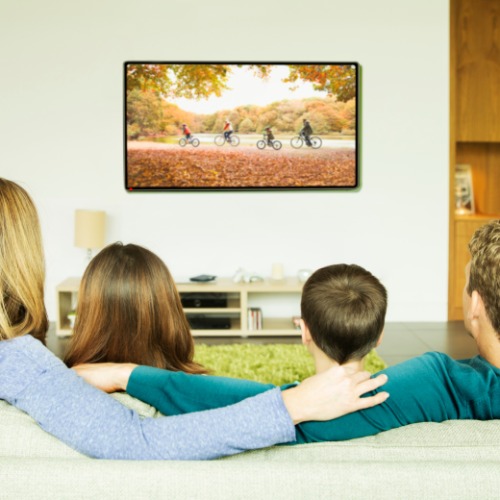 Two adults and two kids are sitting on a sofa while looking at a wall-mounted TV. The living room has a small console.