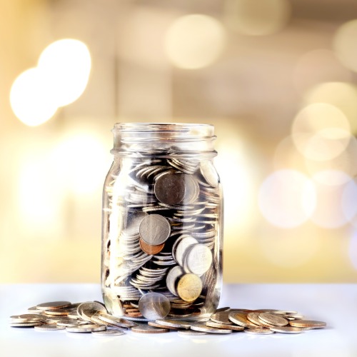 A small Mason jar on a white table. The jar is full of different coins and the coins are spilling out around the jar.