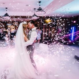 A bride and groom dance together indoors as confetti falls around them, with lights and guests visible in the background.