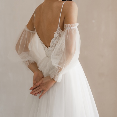 A person wearing a white backless dress with sheer long sleeves, standing with their hands behind back.