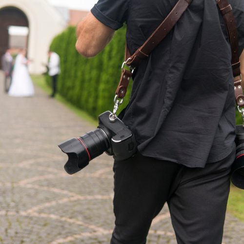 A wedding photographer walking outside with two small cameras toward a newlywed couple and another guest.