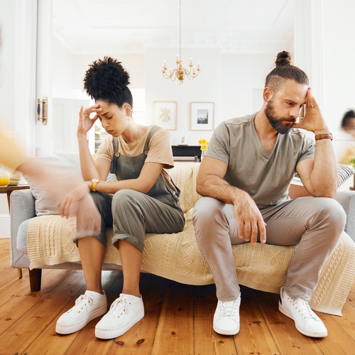 A stressed couple sits on a couch in a bright living room while children run around them, creating a sense of chaos.
