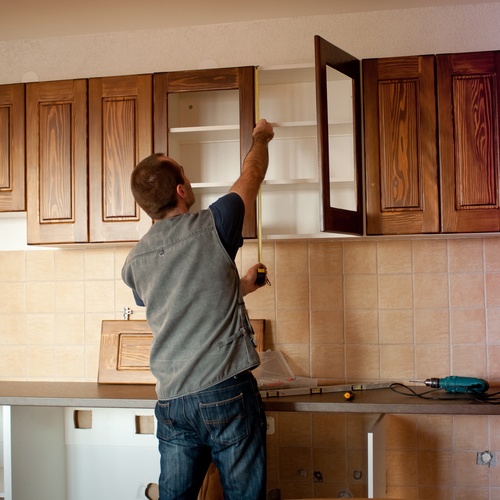 A man in jeans and a vest uses a measuring tape to measure the wood cabinetry in a kitchen under construction.