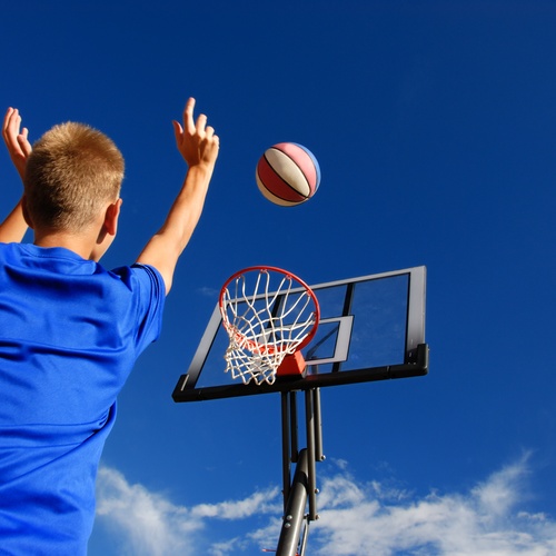 A ground-level view looking up from behind a young boy in a blue t-shirt, shooting a basketball at a hoop.