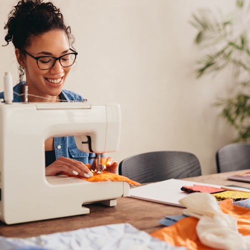 A woman uses a sewing machine. Supplies and pieces of orange, blue, and white fabric are spread across the table.